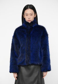 Γυναίκα με λαμπερό μπλε και μαύρο ψεύτικο γούνινο μπουφάν με ψηλό γιακά, συνδυασμένο με μαύρη δερμάτινη φούστα, που στέκεται μπροστά σε λευκό φόντο.