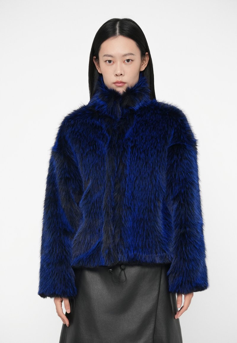 Γυναίκα με λαμπερό μπλε και μαύρο ψεύτικο γούνινο μπουφάν με ψηλό γιακά, συνδυασμένο με μαύρη δερμάτινη φούστα, που στέκεται μπροστά σε λευκό φόντο.