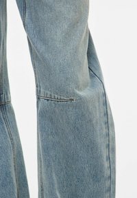 Gros plan d’un jean en denim bleu clair avec des coutures visibles et des détails de couture au niveau du genou légèrement plié sur un fond uni.