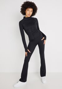 Čierny športový outfit pozostávajúci z priliehavého zipsového saka a rozšírených nohavíc, vyrobený z elastického materiálu. Model má na sebe biele tenisky.