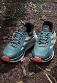 Paar türkisfarbene Laufschuhe mit weißen Streifen und orangen Akzenten, die auf dem Boden zwischen Gras und kleinen Zweigen liegen.