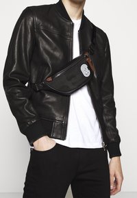 Černá kožená bomber bunda se zipem, pod ní bílý tričko a černé kalhoty. Součástí je černý páskový batoh s hnědými akcenty a logem.