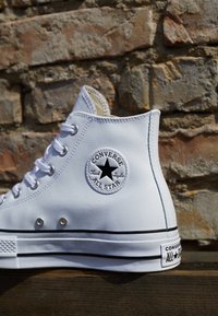Białe sneakersy typu high-top o gładkiej syntetycznej fakturze, z czarnymi akcentami i okrągłym logo po zewnętrznej stronie. Posiadają metalowe oczka i gumową podeszwę.