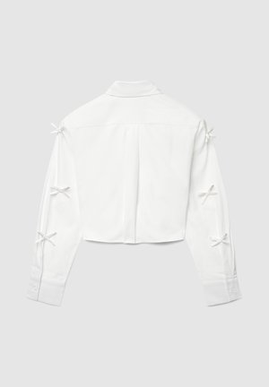 Weißes, langärmeliges, kurzes Hemd mit Kragen und sechs kleinen Schleifen entlang der Rückseite beider Ärmel, auf einem grauen Hintergrund flach ausgelegt.