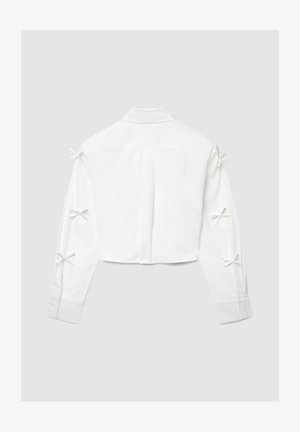 White long-sleeve cropped shirt with collar and six small bows tied along the back of both sleeves, laid flat on a gray background.