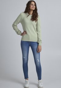 Lys grøn strikket sweater med ribbede manchetter og kant, parret med slidte blå skinny jeans og hvide sneakers, der viser en afslappet stil.