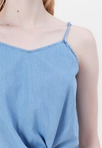 Haut en denim bleu clair avec fines bretelles réglables, doté d'un décolleté en V et d'un détail noué à l'avant. Texture douce et lisse.