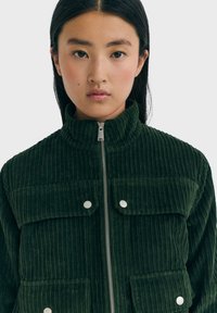 Groene corduroy jas met een hoge kraag, ritssluiting aan de voorkant, twee borstzakken en drukknopdetails, met een geribbelde textuur.