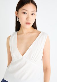 White sleeveless top with a deep V-neck, gathered detail at the bust, and a smooth cotton texture. Model wears a large hoop earring.