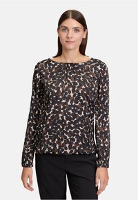 Frau mit glattem braunem Haar trägt eine langärmlige Bluse mit Leopardenmuster und schwarze Hose und steht vor einem schlichten hellen Hintergrund.