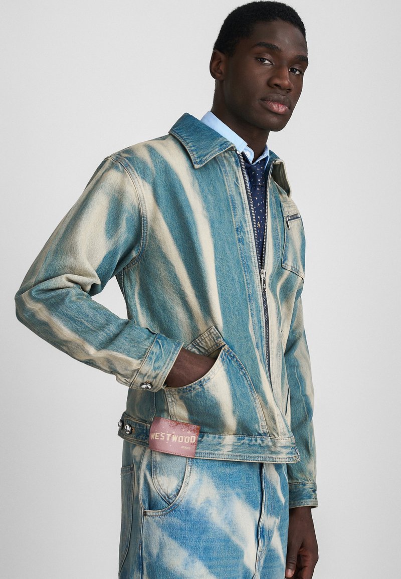 Giacca di jeans con un motivo tie-dye in blu e beige, con chiusura a zip, due tasche laterali e un'etichetta rosa sul retro.