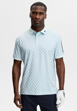 Man draagt lichtblauw poloshirt met patroon en korte mouwen, met witte golfhandschoen aan rechterhand, staand tegen een effen achtergrond.