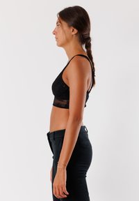 Bralette in pizzo nero con spalline regolabili, texture trasparente e design aderente. Abbinato a pantaloni neri a vita alta. Vista laterale.