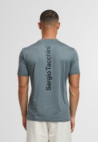 Grå kortærmet træningsshirt med en glat tekstur. Navnet "Sergio Tacchini" fremgår lodret med fede sorte bogstaver på bagsiden.