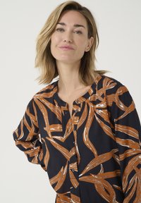 Chemise noire avec un motif de feuilles orange et blanches sur l'ensemble, dotée d'un col rond, d'une fermeture à boutons et de manches longues avec une coupe décontractée.