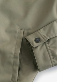 Tela de chaqueta verde oliva con una textura suave, que presenta puños abotonados y una abertura lateral para un detalle de ventilación.