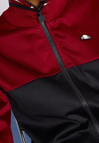 Rot schwarze Zip-Jacke mit hohem Kragen, mit glatter Textur, einem Logo auf der Brust und kontrastierenden Farbbereichen.