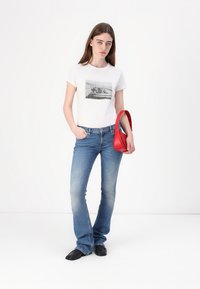 T-shirt en coton blanc avec un imprimé graphique, assorti à un jean évasé bleu clair. Tient un petit sac à main rouge et porte des chaussures noires.