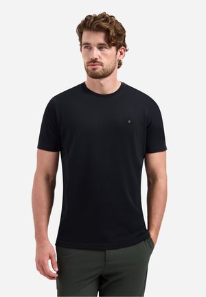 Man met krullend haar en baard, gekleed in een effen zwart T-shirt met korte mouwen en donkergroene broek, staand met één hand in de zak.