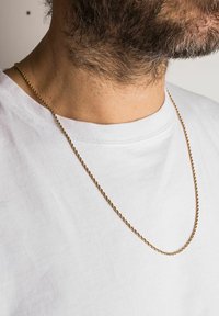 Guldtonet snoet kæde halskæde båret over en hvid t-shirt, der fremhæver dens glatte tekstur og reflekterende overflade mod huden.
