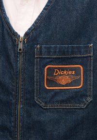Μπουφάν από σκούρο μπλε τζιν με πορτοκαλί ραφές στην μπροστινή τσέπη, διακοσμημένη με ένα μπατς λογότυπο Dickies Harley-Davidson και κεντρικό μεταλλικό φερμουάρ.