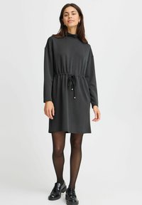Egy nő fekete, hosszú ujjú ruhában, húzózsinóros derékkal, fekete áttetsző harisnyában és fekete mokaszin cipőben áll egy fehér háttér előtt.