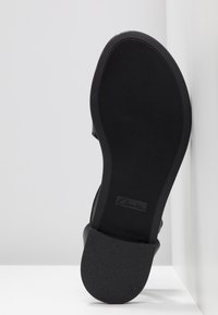 Semelle de chaussure noire avec un motif fin de bande de roulement horizontale et une zone rectangulaire pour le logo, positionnée verticalement contre une surface blanche.