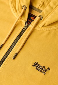 Sudadera con cremallera amarilla de tejido de algodón texturizado, con cremallera oscura, cordones ajustables y logo bordado "SuperDry".