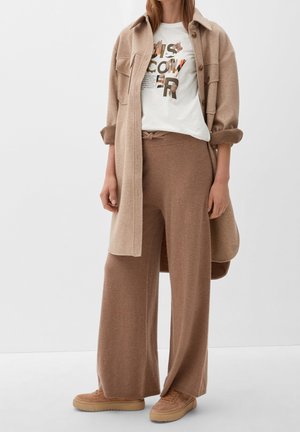 Beige Wolle-Blend langes Hemd, weißes Grafik-T-Shirt mit Blätterdesign, hellbraune weit geschnittene Hose und passende beige Sneakers.