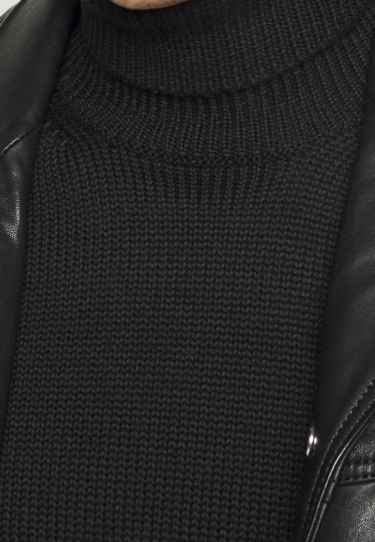 Gros plan sur un pull noir à col roulé en tricot porté sous une veste en cuir noire avec un bouton-pression en argent visible.