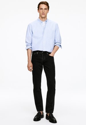 Jasnoniebieska w biało-szare paski koszula z guzikiem, czarne dżinsy i czarne mokasyny. Koszula ma kieszonkę na piersi i podwinięte rękawy.