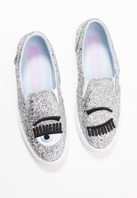 Silvriga glittriga slip-on skor med broderade blinkande och  öppna blå ögonmönster, ljusblå insida och vita sulor.