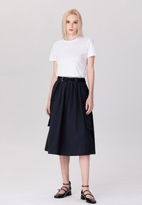T-shirt blanc associé à une jupe midi noire marine taille haute avec une ceinture, des poches et un design fluide. Des chaussures à brides noires complètent le look.