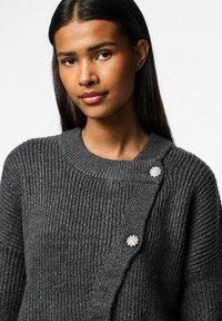 Pilkas mezgimo megztinis su banguota tekstūra, apvaliu iškirpimu ir šoniniu užsegimu, kuriame yra du dekoratyviniai kristaliniai gėlių mygtukai.