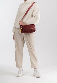 Czerwona nylonowa torba na ramię z zamykanymi kieszeniami, żółtym frędzlem i haftowanym logo. Nosi się z beżowymi spodniami w luźnym kroju i białymi sneakersami.