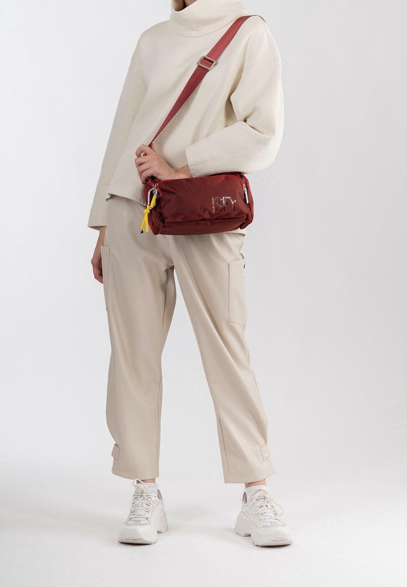 Czerwona nylonowa torba na ramię z zamykanymi kieszeniami, żółtym frędzlem i haftowanym logo. Nosi się z beżowymi spodniami w luźnym kroju i białymi sneakersami.