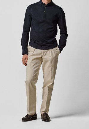 Langærmet mørkeblå polo med tre-knapslukning, kombineret med lys beige plisserede bukser og mørkebrune loafers.