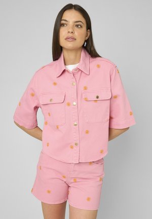Femme portant une chemise rose à manches courtes boutonnée et un short assorti avec de petits imprimés de fleurs orange, debout les mains dans le dos.