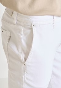 Pantalones de mezclilla blanca con un acabado texturizado. Presenta bolsillos laterales, herrajes plateados y una etiqueta en la cintura de color beige claro.