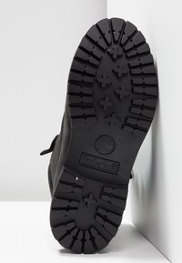 Semelle de botte noire avec des crampons profonds, motifs d'adhérence en forme de croix, logo Timberland, photographiée en position verticale sur fond blanc.