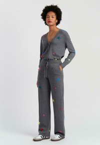 Grijze gebreide jumpsuit met kleurrijke sterrenpatronen, voorzien van een V-hals, lange mouwen en een trekkoord in de taille. Gepaarde met wit-grijze sneakers.