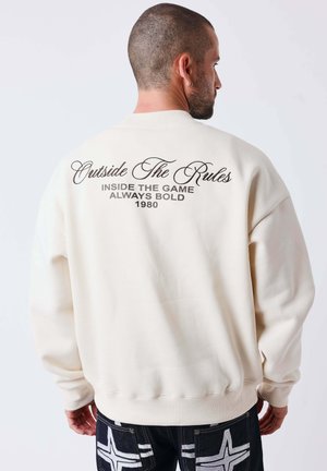 Homme avec la tête rasée portant un sweat-shirt blanc avec le texte "Outside The Rules, Inside The Game, Always Bold 1980" dans le dos.