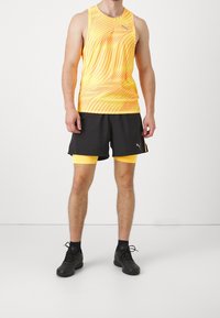 Puma sportovní oblečení s žlutým tílkem bez rukávů a vlnitými oranžovými vzory, kombinované s černými šortkami a žlutými akcenty. Černé atletické boty.