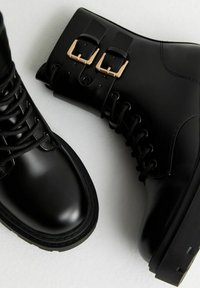 Botas de tobillo de cuero negro con punta redonda, cordones en la parte frontal y dos hebillas como acentos. Cuenta con una suela de goma negra resistente y un acabado suave.