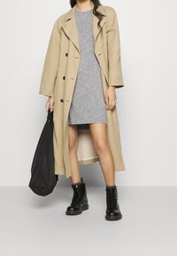Trench coat beige con grandi bottoni, indossato sopra un vestito grigio aderente. Stivali neri stringati e una borsa nera completano l'outfit.