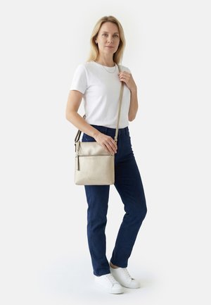 Frau mit schulterlangem blondem Haar, trägt weißes T-Shirt, blaue Jeans, weiße Sneakers und hält eine beiger Umhängetasche über der Schulter.