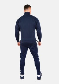 Temno-modra zip-jemna jakna in usklajene hlače iz gladkega materiala. Ima visok ovratnik in prilegajoč design, obležena z črno-belimi supergami.