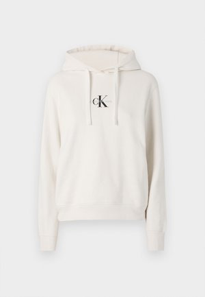Mikina s kapucňou v jemne bielej farbe, so predným logom s textom "CK". Vyrobená z mäkkej tkaniny s voľným strihom a rebrovanými manžetami.