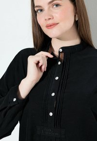 Blusa in corduroy nera con colletto alto, patta dei bottoni e dettagli texturizzati sottili. Presenta una cucitura decorativa marrone vicino al collo.