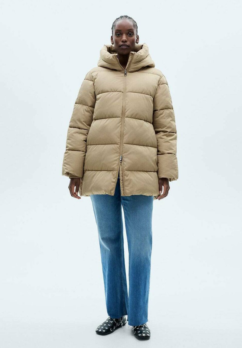 Beige pufferjack met capuchon, voorzien van doorgestikte panelen en een ritssluiting aan de voorkant. Gekleed met lichtblauwe straight-leg jeans en zwarte patroon schoenen.
